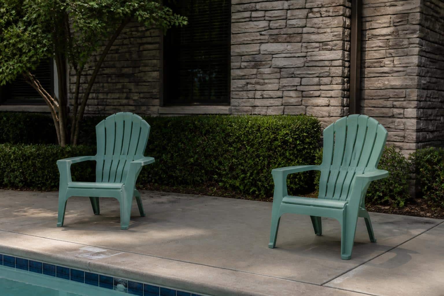 Two seafoam green plastic Adirondack chairs spaced well apart on a concrete pool deck near the water edge