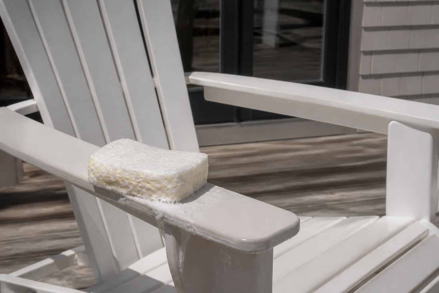 A soapy sponge wiping the wide flat armrest of a white plastic Adirondack chair, suds on the smooth surface