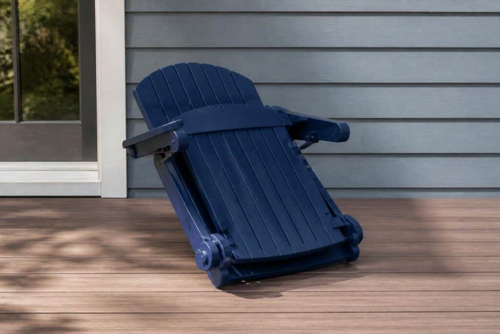 A navy blue folding plastic Adirondack chair shown partially folded flat on a deck, folding hinge visible