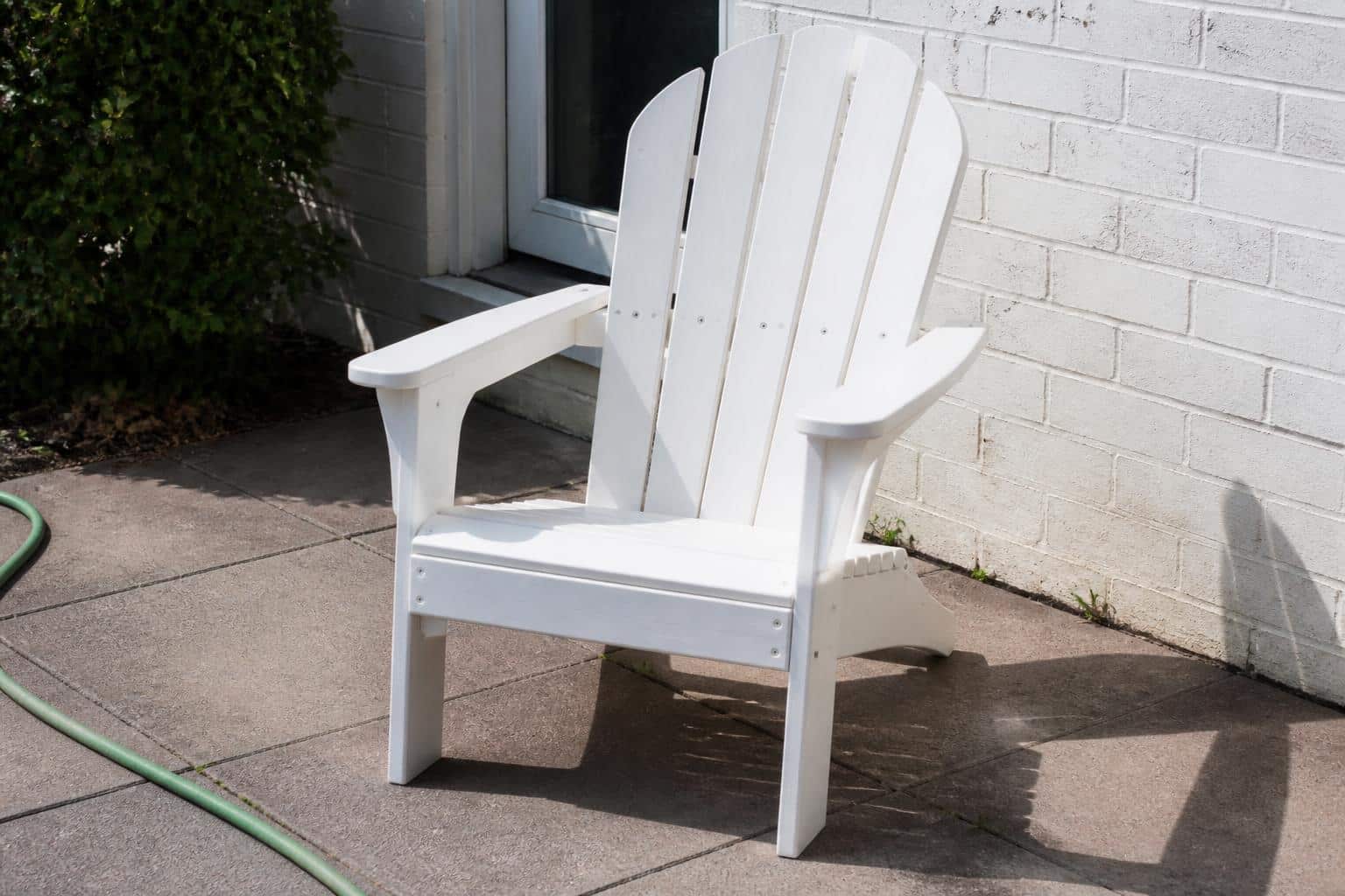 Documentary-style phone photo of a well-kept bright white plastic patio chair that has clearly spent years outdoors on a lived-in patio with a garden hose nearby