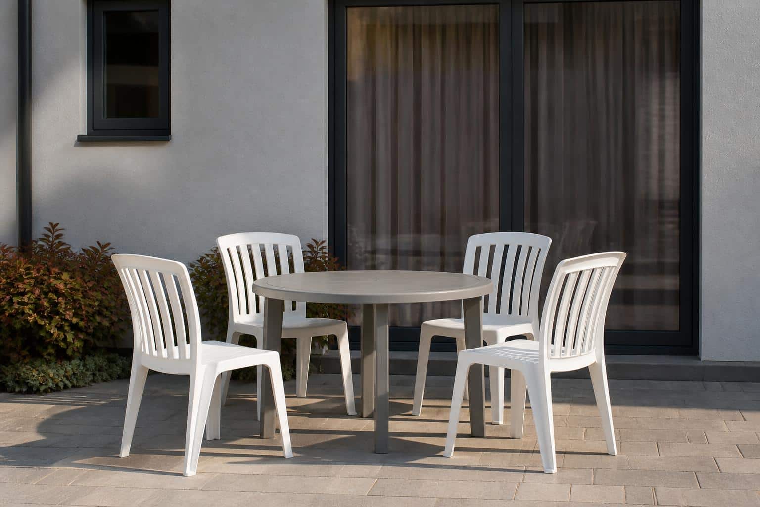 Four white armless plastic dining chairs evenly spaced around a round gray plastic patio table on clean pavers, crisp clean edges and shadows