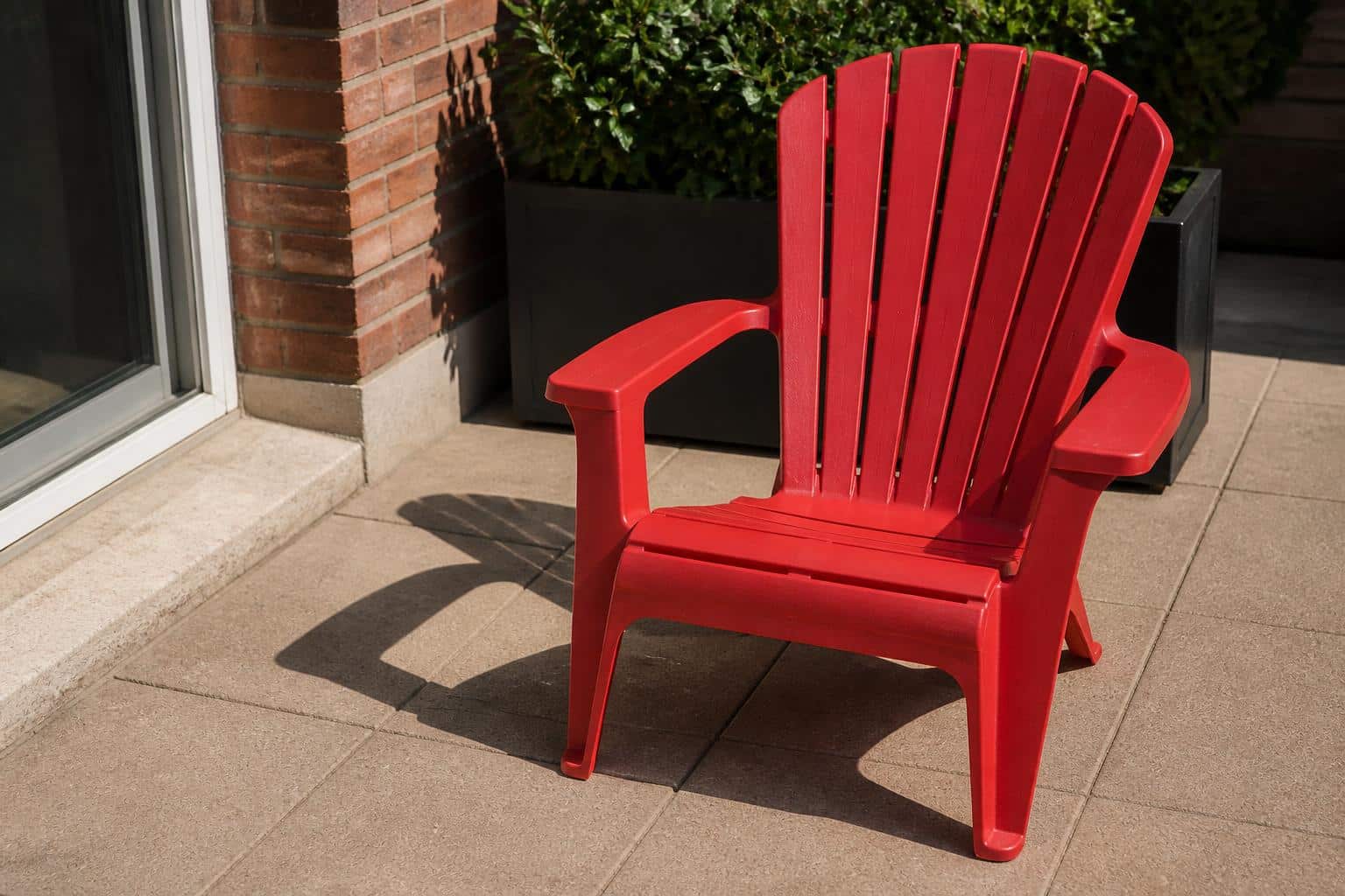 A bright red plastic Adirondack chair in strong afternoon sun casting long shadows on a patio
