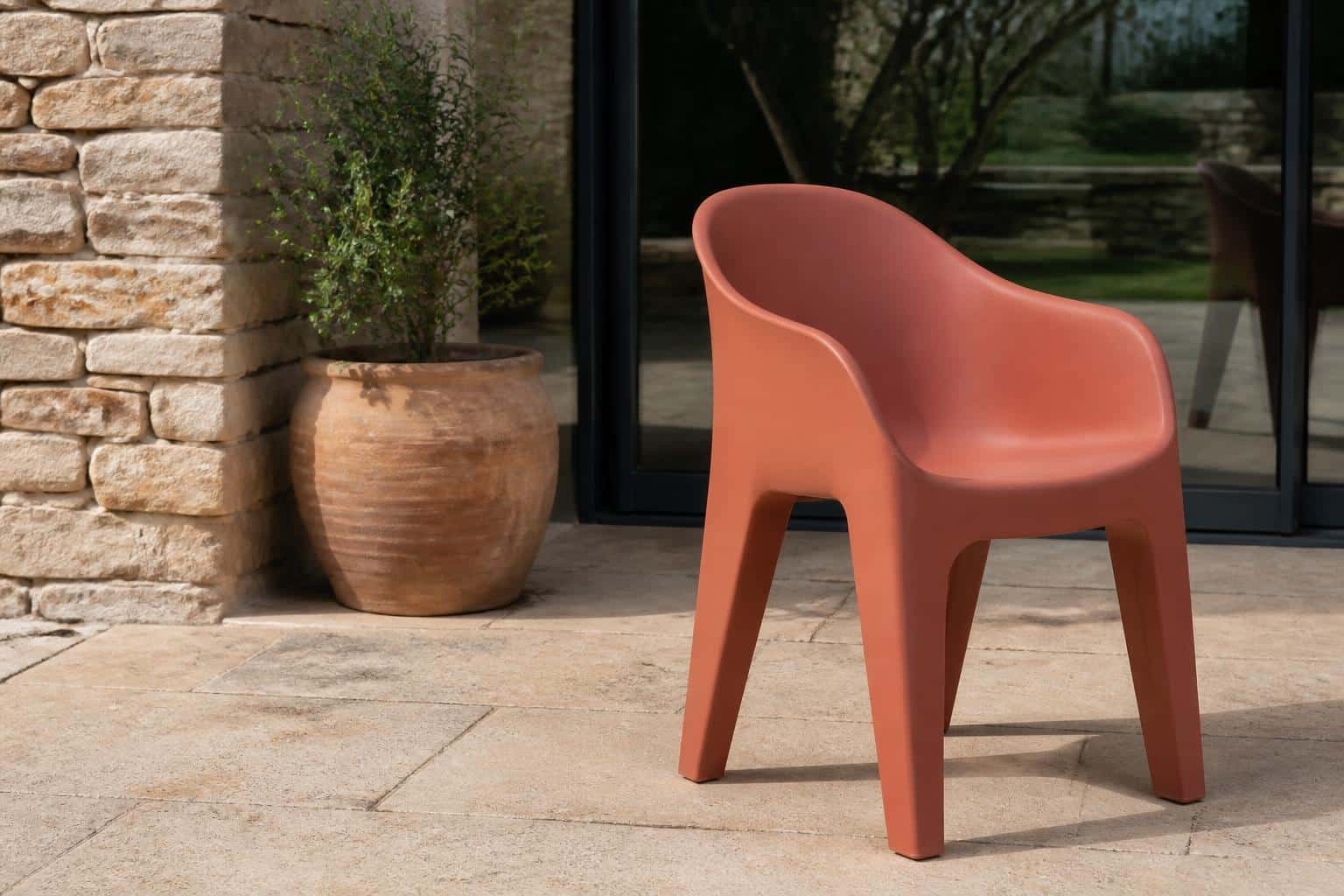 A one-piece smooth molded resin patio chair in terracotta with a seamless curved shell form, no slats, on a stone patio